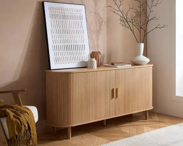 A light natural wood sideboard cabinet with vertical fluting and two small gold-toned handles, styled in a warm beige room with a framed print, white vase, and branch decor.