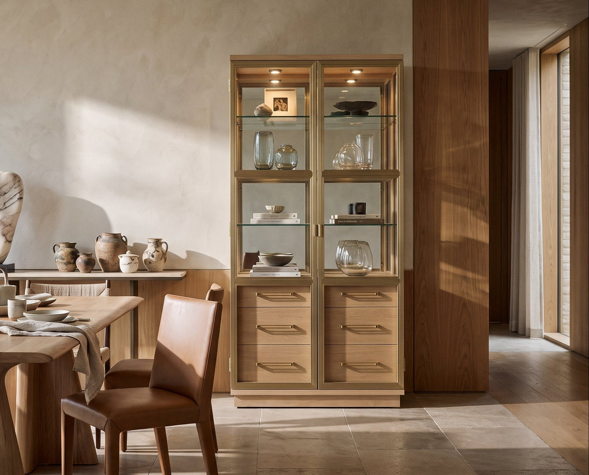 A tall wooden display cabinet with glass shelves stands in a sunlit dining room beside a wooden table and chairs. The cabinet has warm wood drawers and upper glass cabinets, and the room has textured plaster walls and large windows.
