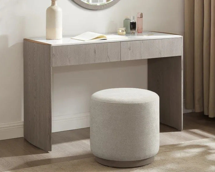 A modern vanity desk with a light gray wood-grain finish and a white top, paired with a matching cylindrical upholstered stool in a bright room.