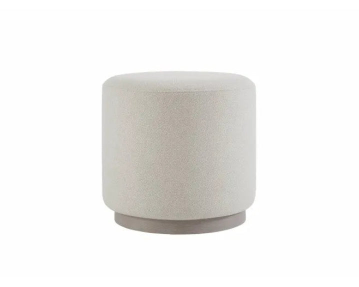 A cylindrical upholstered ottoman or stool in a light warm gray or off-white fabric, shown against a plain white background.