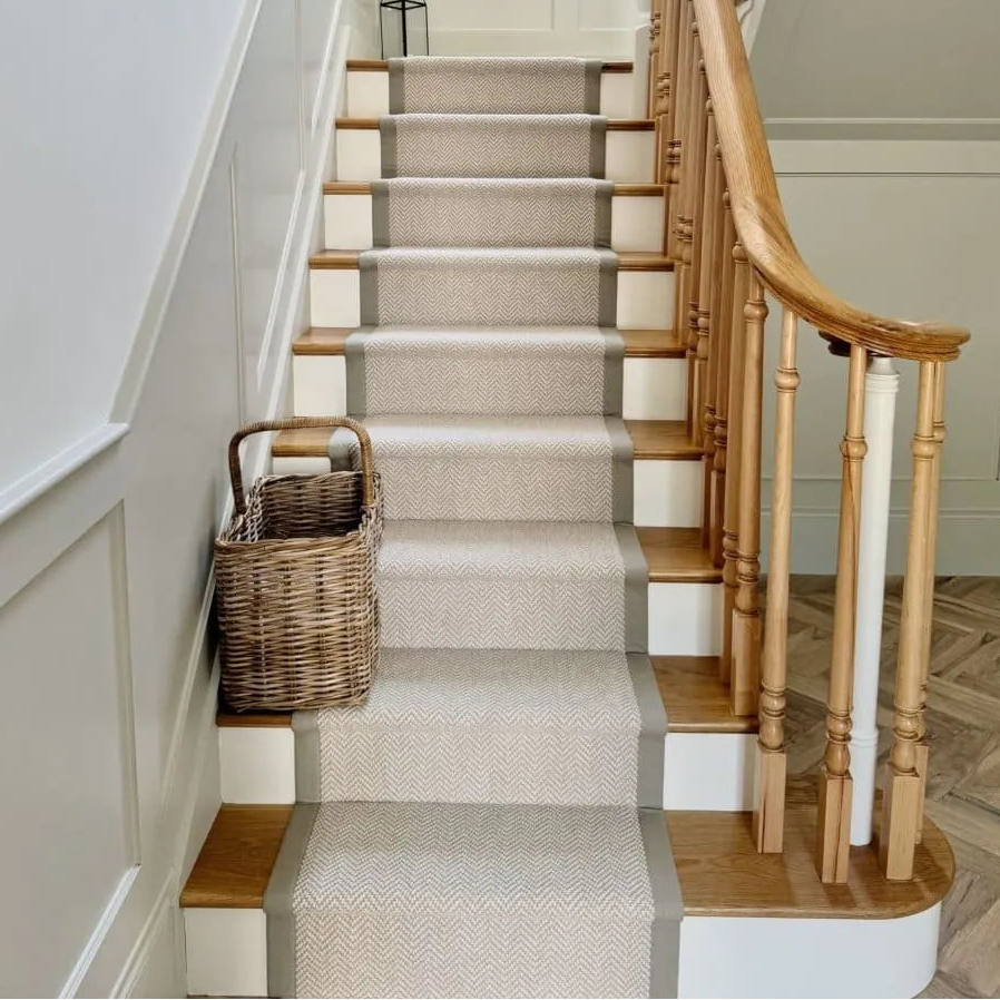 A carpet stair runner in a light beige textured fabric with gray borders, installed on a wooden staircase with white risers. A wicker basket sits on the landing next to the stairs, and a black lantern hangs at the top.