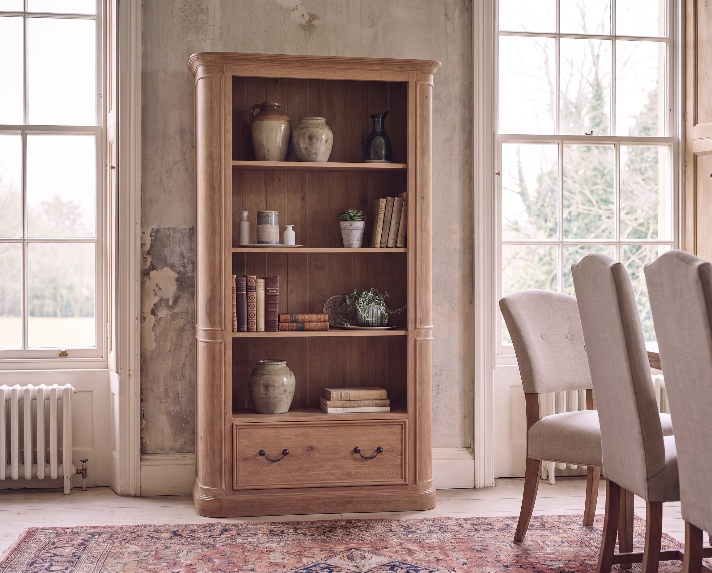 A tall wooden bookcase with three shelves and a single drawer, styled with books and decorative vases in a rustic room with large windows and dining chairs.