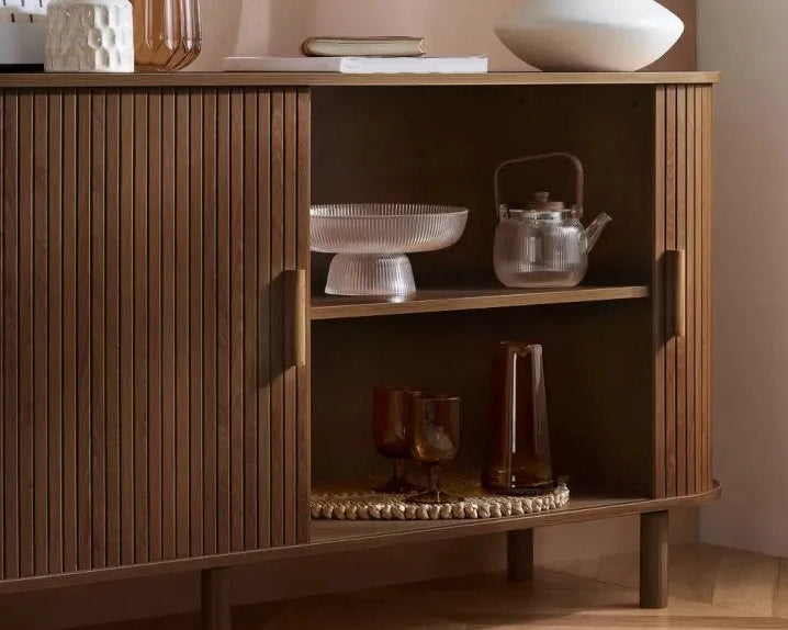 A mid-century modern wooden sideboard cabinet with vertical fluted doors, open shelving, and brass-toned handles, styled with glassware and a teapot in a warm interior setting.
