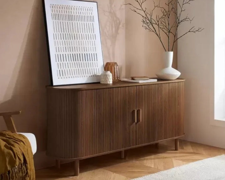 A mid-century modern wooden sideboard cabinet with vertical fluting, styled in a warm beige room with a framed art print, white ceramic vase, and decorative objects.