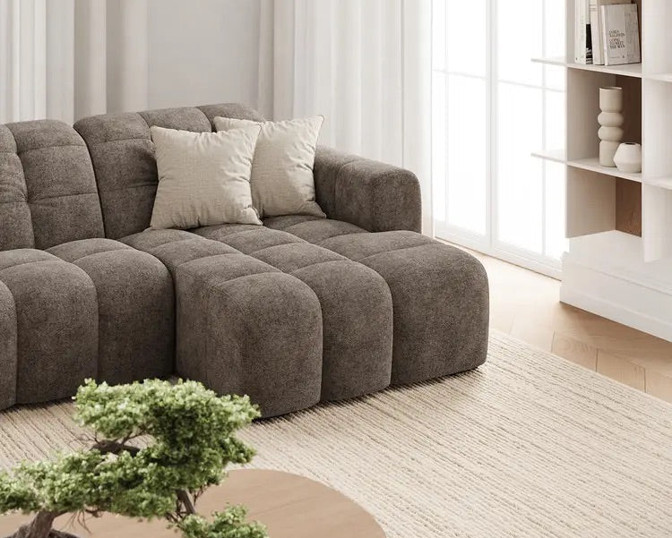 A modern modular sectional sofa in textured taupe-brown fabric, styled in a bright, minimalist living room with white pendant lights, a bookshelf, and a woven rug.