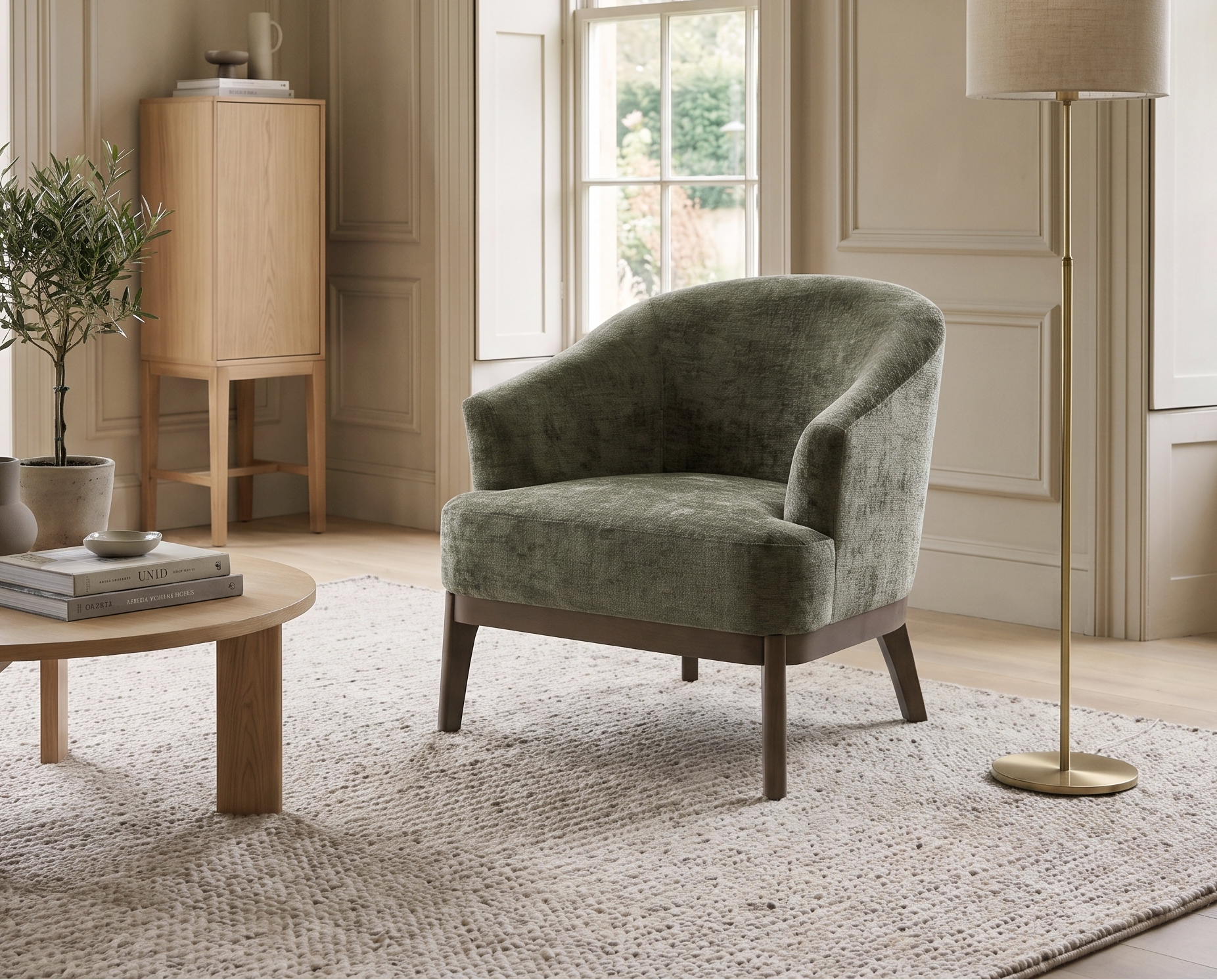 A green velvet armchair in a beautifully styled, sunlit room with beige walls, a round wooden coffee table, a floor lamp, and a textured rug.