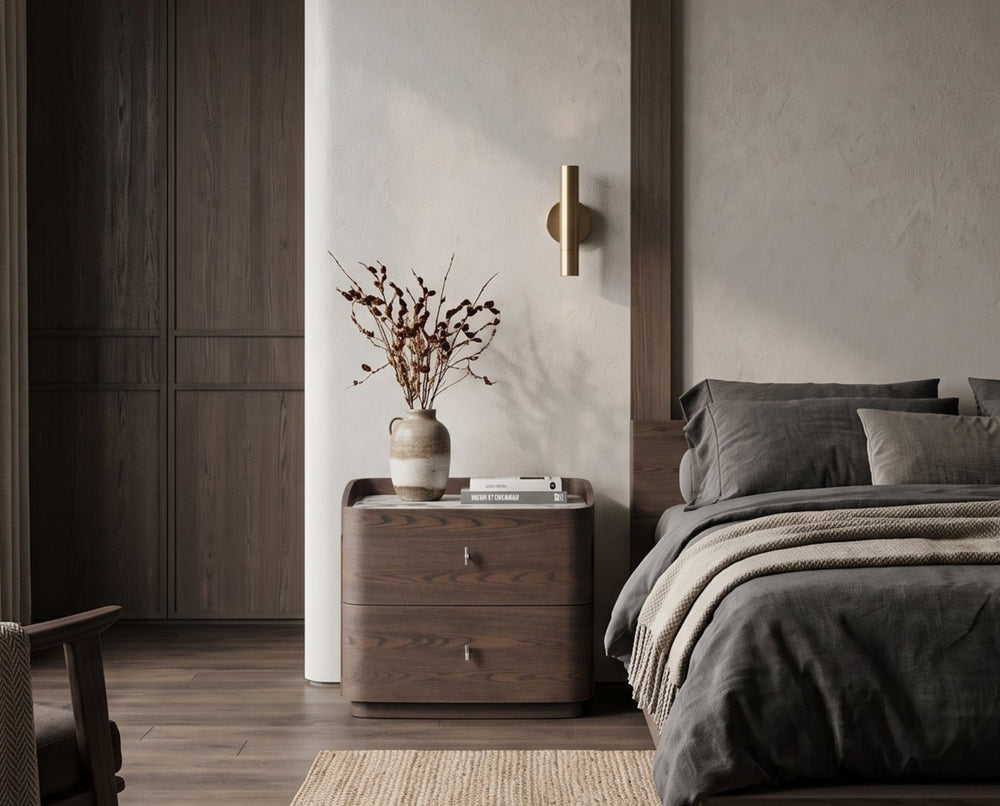 A modern bedroom interior featuring a bed dressed in dark charcoal bedding, a textured beige rug, a wooden nightstand with a vase of branches, and a dark armchair near a large window.