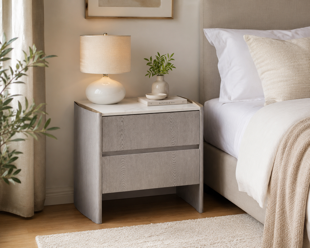 A modern two-drawer nightstand in a warm gray wood-grain finish, styled in a bedroom next to a bed with white linens, a white table lamp, and a small potted plant.