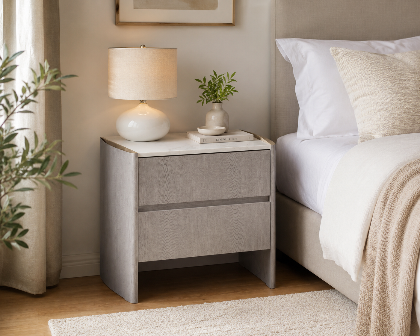 A modern two-drawer nightstand in a warm gray wood-grain finish, styled in a bedroom next to a bed with white linens, a white table lamp, and a small potted plant.