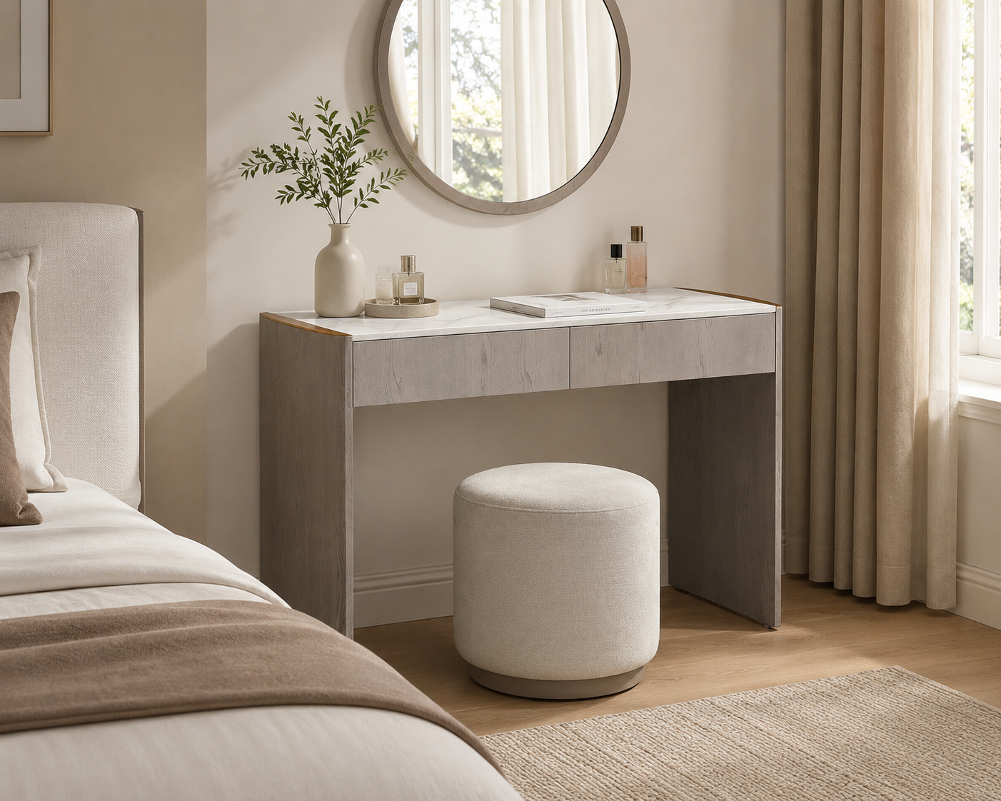 A modern vanity desk with a light gray marble top and textured gray side panels, paired with a matching cylindrical ottoman in a softly lit bedroom. A round wall mirror, a beige vase with greenery, and small decorative bottles sit on the desk.