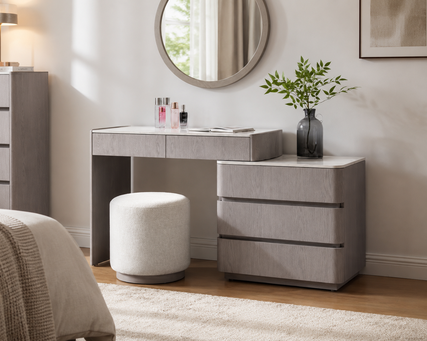 A modern gray vanity table set in a bright bedroom, featuring a matching stool, a round wall mirror, and a dark glass vase with greenery on top.