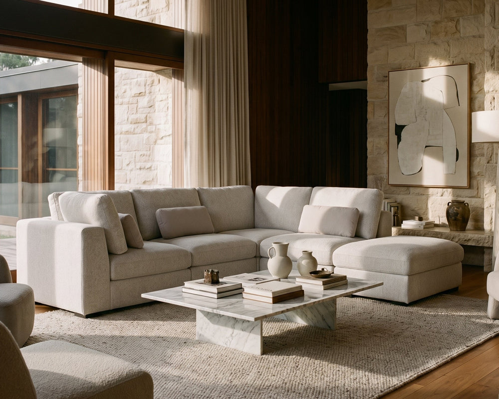 A modern living room featuring a light beige sectional sofa with matching armchairs, a marble coffee table, a textured area rug, and a stone wall with abstract wall art.