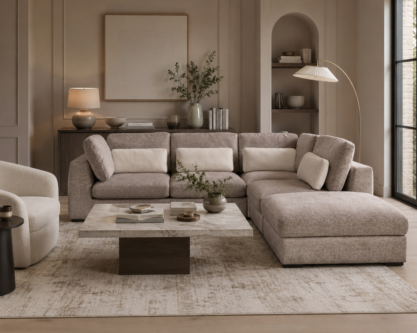 A modern sectional sofa in a warm taupe fabric with matching throw pillows, styled in a contemporary living room with a marble-top coffee table, textured rug, and a matching armchair.
