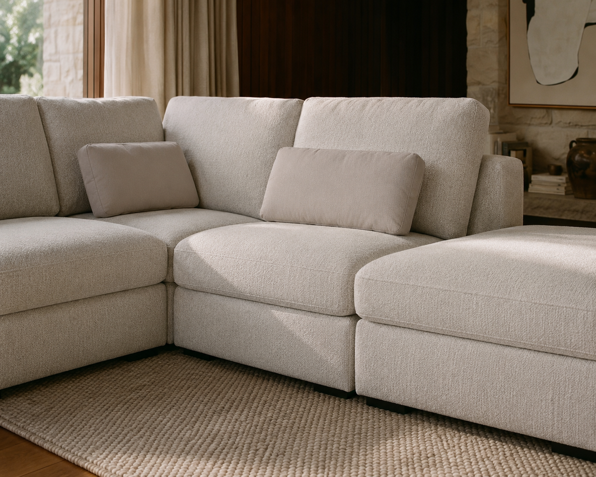 A light beige upholstered sectional sofa with two matching throw pillows, shown in a styled living room setting with a woven rug, curtains, and a stone wall.