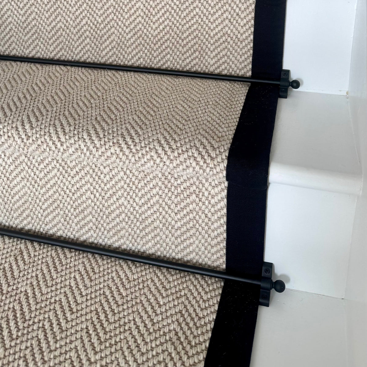 A close-up view of a carpet stair runner installed on a staircase. The runner features a beige and brown herringbone pattern with solid black side borders and black metal railings.