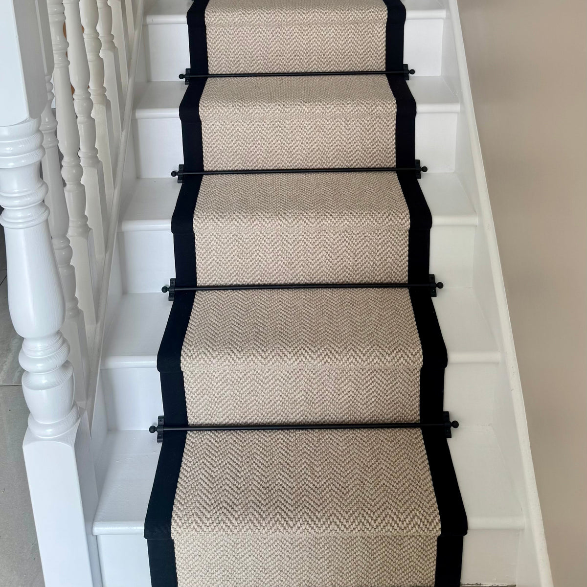 A beige and brown herringbone stair runner with black borders and black metal rails installed on a white staircase.