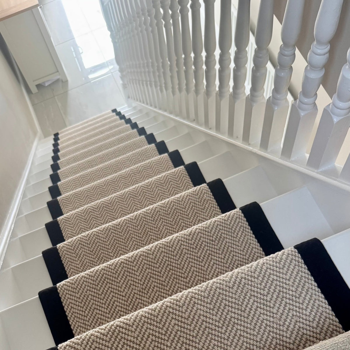 A set of beige and brown herringbone stair carpet runners installed on a staircase with white risers and a white wooden banister.