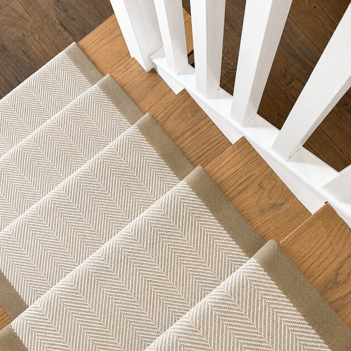 A close-up view of carpeted stair treads with a beige and white herringbone pattern, installed on wooden stairs next to a white wooden railing.