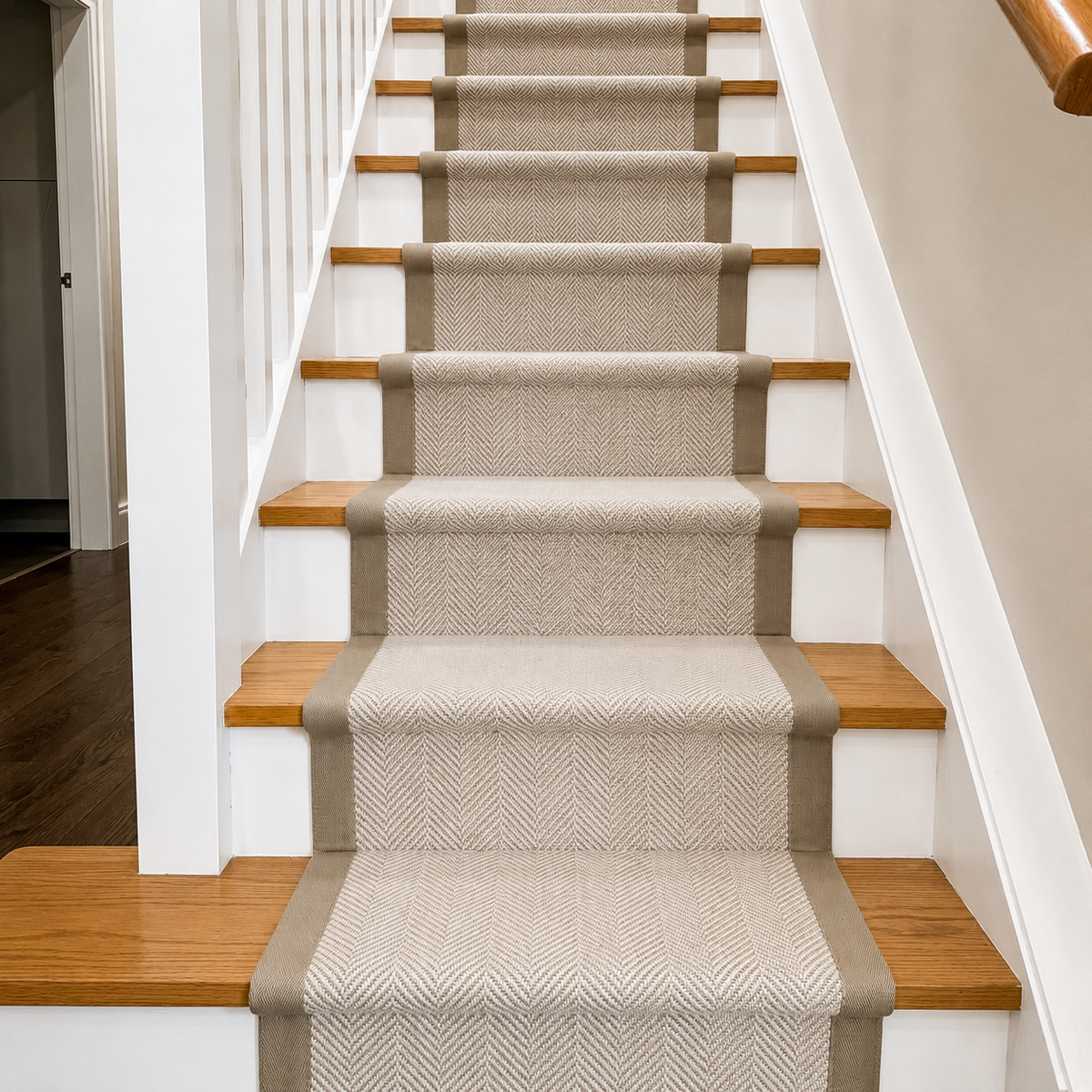 A set of carpet stair runners with a beige and white herringbone pattern and taupe borders, shown installed on a wooden staircase with white balusters.