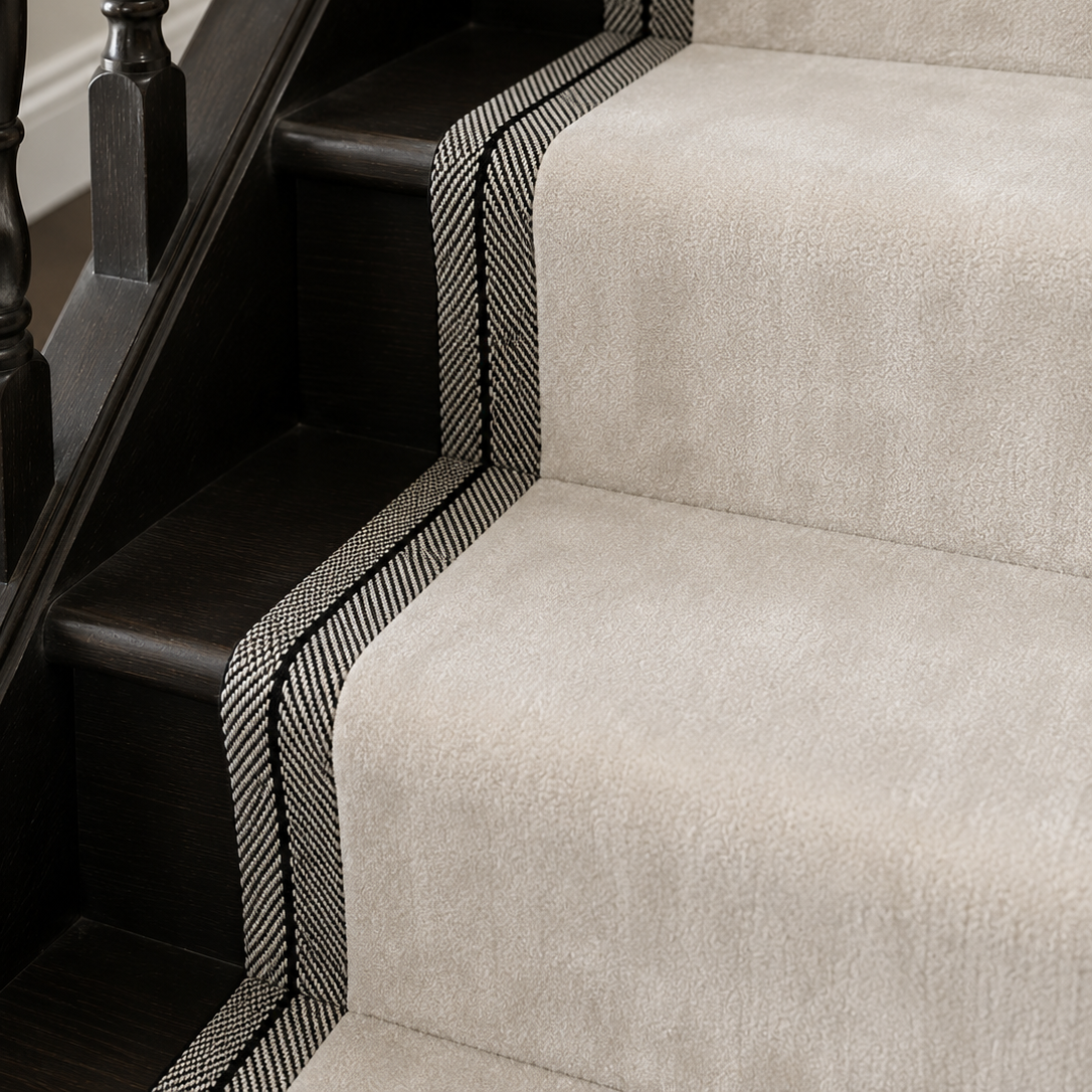 A close-up of carpeted stairs with a light beige stair runner bordered by black and white herringbone patterned trim, next to a dark wood banister.