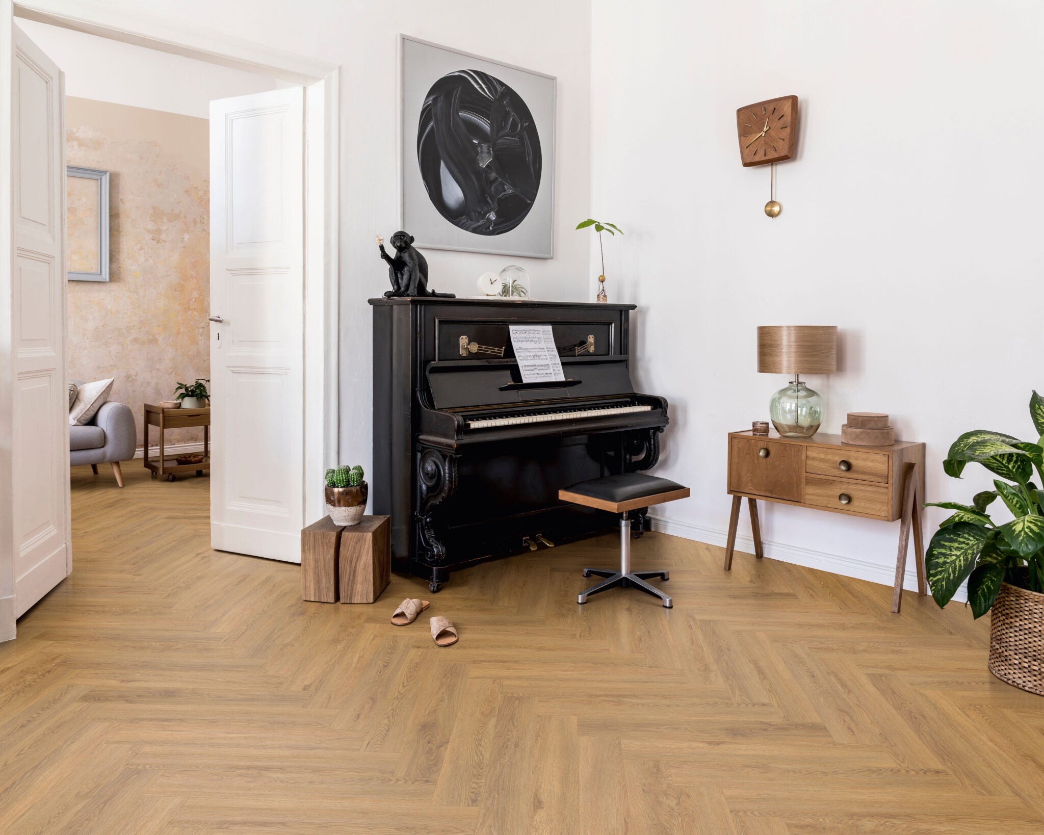 A styled interior room with light oak herringbone wood flooring, featuring a black upright piano, a wooden side cabinet with a lamp, a potted plant, and framed wall art.