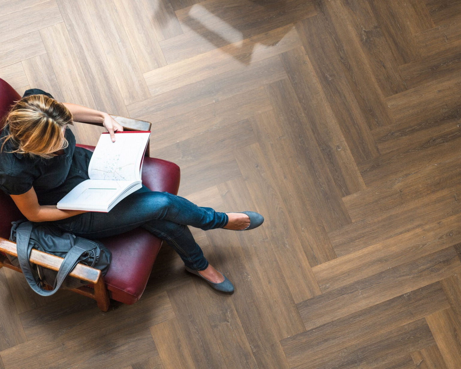 A woman sits in a red armchair reading a book on a dark wood herringbone floor, with a black backpack beside her.