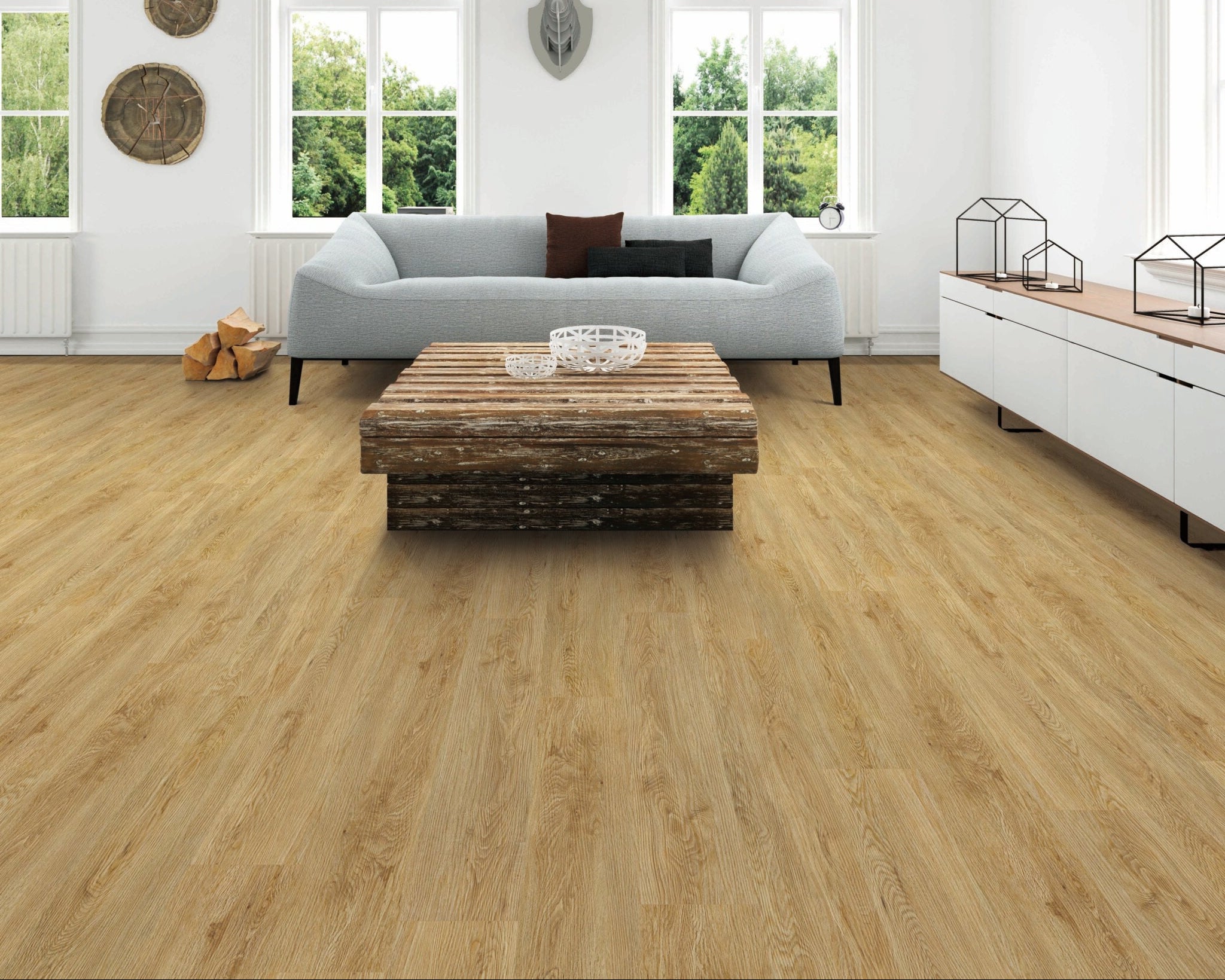 A bright, modern living room with light oak wood plank flooring, featuring a light blue sofa, a rustic wooden coffee table, and white walls with large windows.