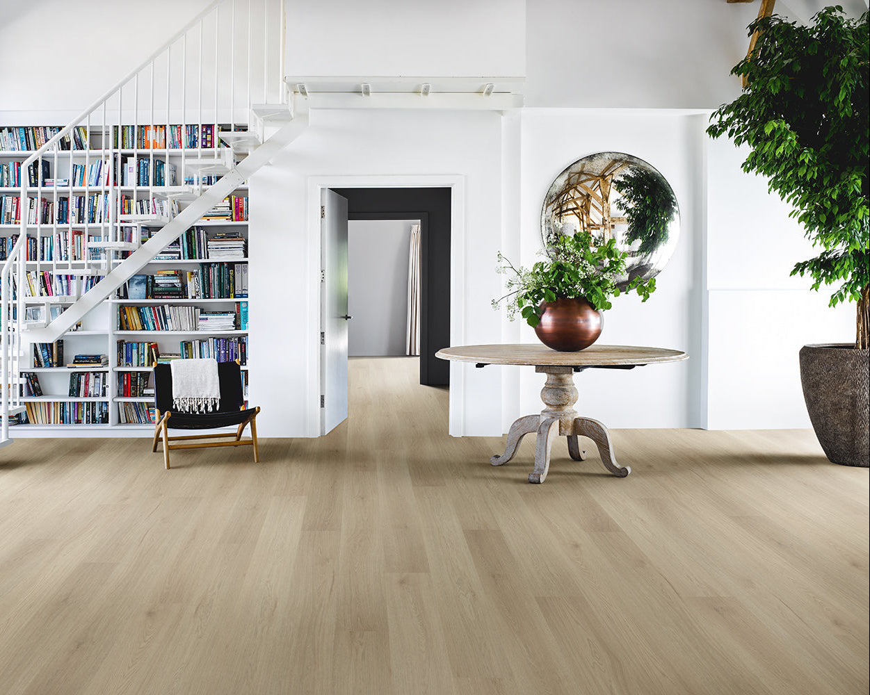 Light oak wood flooring in a bright, modern open-plan living space with a spiral staircase, bookshelves, a round table with a potted plant, and a large green tree on the right.