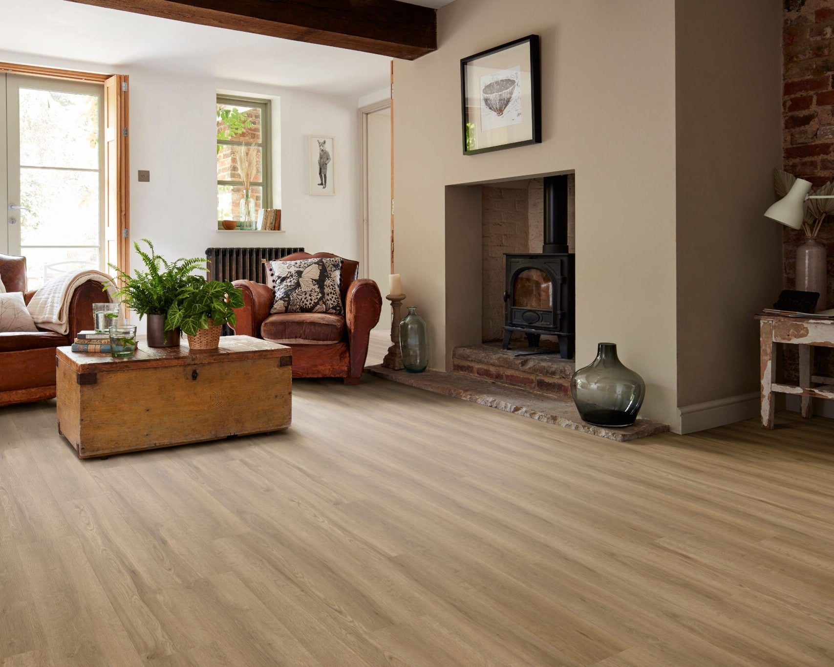 Light oak wood flooring installed in a cozy, well-lit living room with brown leather armchairs, a rustic wooden coffee table, a brick fireplace, and neutral beige walls.